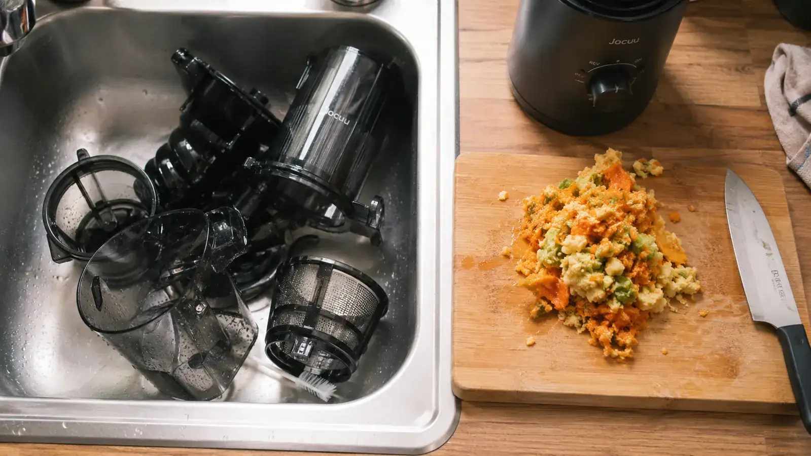 Pile of wet pulp and vegetable scraps on a cutting board next to the disassembled Jocuu juicer parts in a sink
