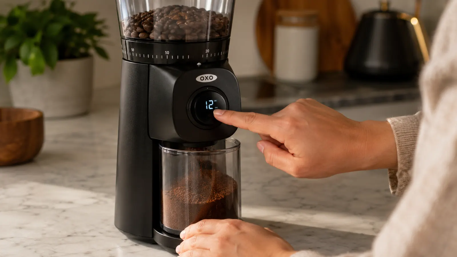Hand pressing the one-touch button on the OXO Brew Conical Burr Grinder while grounds fall into the glass bin below