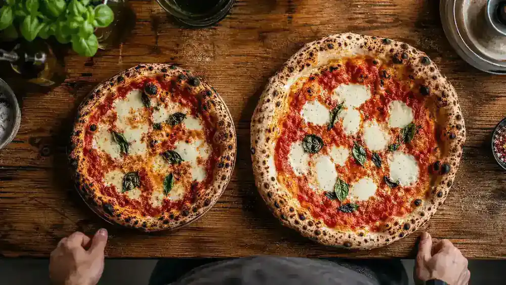 Two pizzas side by side on a wooden table, one from a compact countertop pizza oven and one from a larger freestanding pizza oven, comparing crust size and char