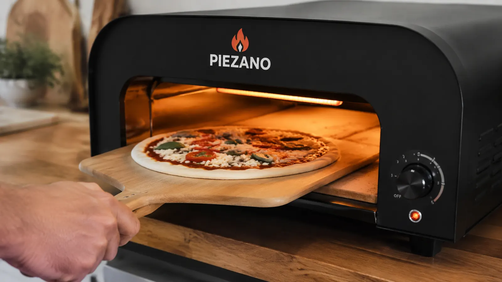 Hand sliding a raw pizza on a peel into the open PIEZANO pizza oven, glowing ceramic stone visible inside